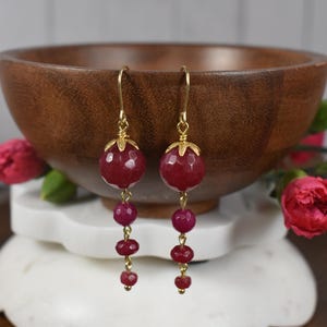 May include: Dangling earrings with gold-colored hooks and accents. Each earring features a large, faceted, deep red bead at the top, followed by smaller, round, red beads. The earrings are displayed in front of a wooden bowl.