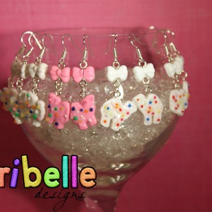 May include: A collection of handmade earrings displayed in a glass. The earrings feature various designs, including white bows, pink bows, and animal shapes, all adorned with colorful sprinkles. The background is a vibrant pink.