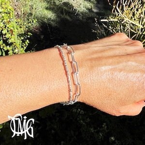 May include: Two silver bracelets on a wrist. One bracelet is a delicate chain with small beads, the other is a thicker chain with larger links.