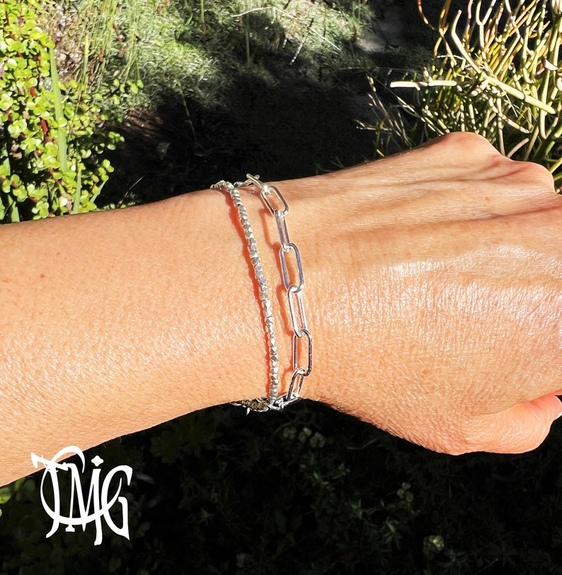 May include: Two silver bracelets on a wrist. One bracelet is a delicate chain with small beads, the other is a thicker chain with larger links.
