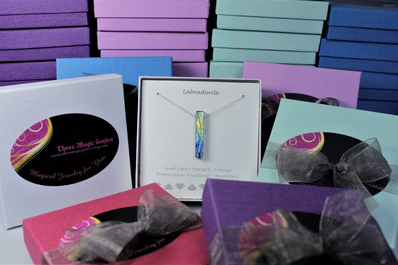 May include: A silver necklace with a blue, green and yellow labradorite bar pendant. The necklace is in a white box with the text "Labradorite" and "Good Luck, Strength, Courage, Perseverance, Confidence, Serendipity". The box is surrounded by other boxes in various colours.