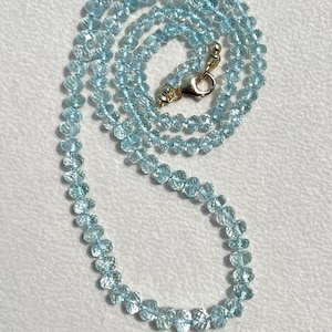 Op de afbeelding: Een delicate ketting bestaande uit kleine, gefacetteerde, lichtblauwe edelstenen. De kralen zijn aan elkaar geregen en vormen een sierlijke curve. De ketting is vastgezet met een goudkleurige sluiting. De achtergrond is zacht, gebroken wit.