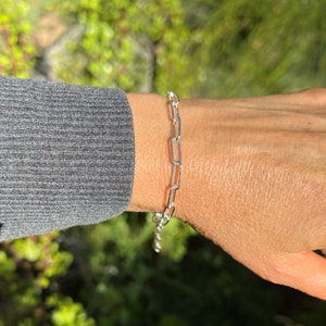 May include: A silver chain bracelet with a simple, linked design. The bracelet is worn on a person's wrist.