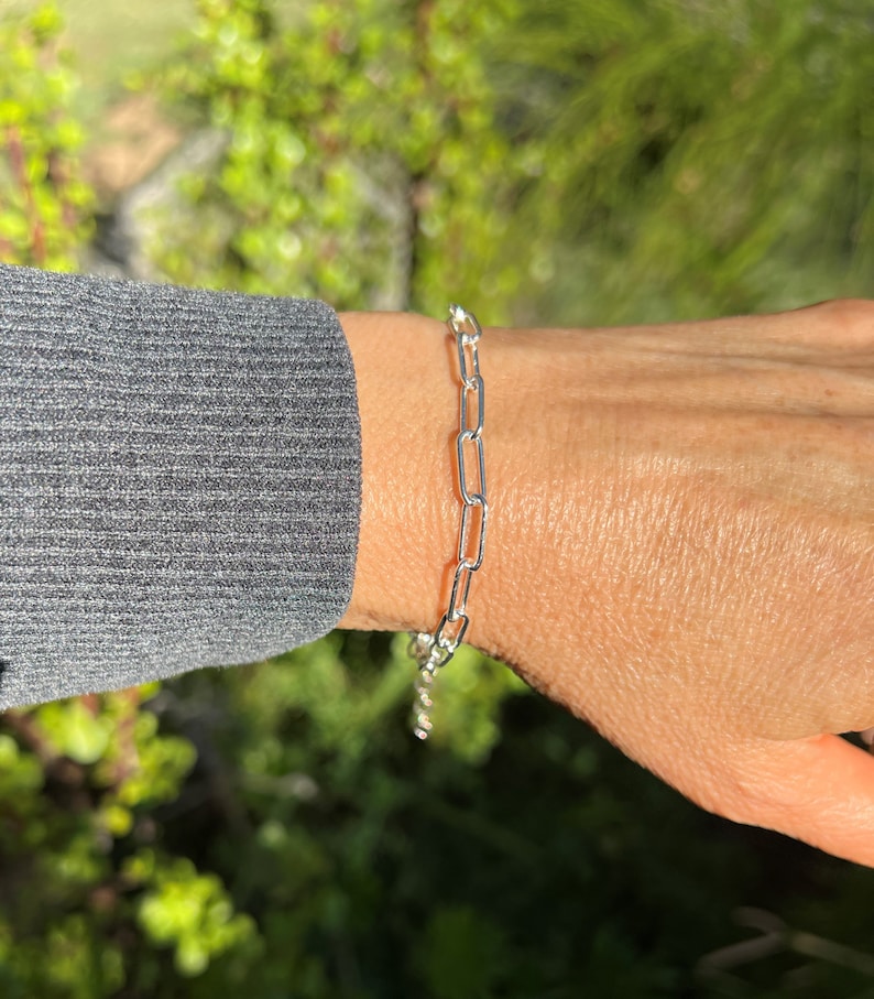 May include: A silver chain bracelet with a simple, linked design. The bracelet is worn on a person's wrist.