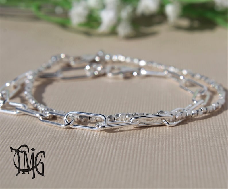 May include: A silver bracelet featuring three different chain styles. The bracelet includes a delicate beaded chain, a thicker chain with oval links, and a chain with small, square links.