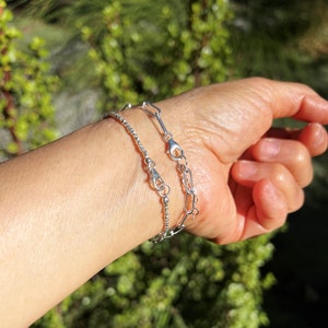May include: Two silver bracelets on a person's wrist. The bracelet on top is a delicate chain with a small clasp. The bracelet on the bottom is a thicker chain with a larger clasp.
