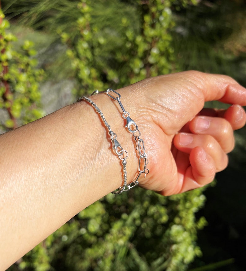 May include: Two silver bracelets on a person's wrist. The bracelet on top is a delicate chain with a small clasp. The bracelet on the bottom is a thicker chain with a larger clasp.