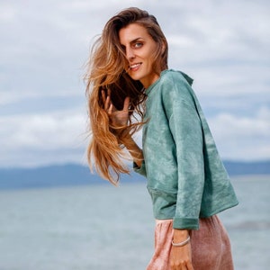 May include: A woman with long brown hair wearing a light green tie-dye sweatshirt and a pink skirt. She is standing in front of a body of water with a cloudy sky in the background.