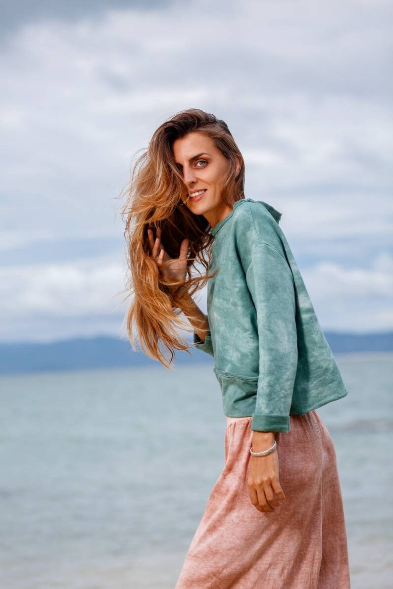 May include: A woman with long brown hair wearing a light green tie-dye sweatshirt and a pink skirt. She is standing in front of a body of water with a cloudy sky in the background.