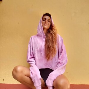 K&ouml;nnte beinhalten: Ein hellrosa Tie-Dye-Hoodie mit Kapuze und Kordelzug sowie langen &Auml;rmeln. Das verk&uuml;rzte Sweatshirt wird mit schwarzen Shorts kombiniert. Das Model hockt vor einer gelben Wand.