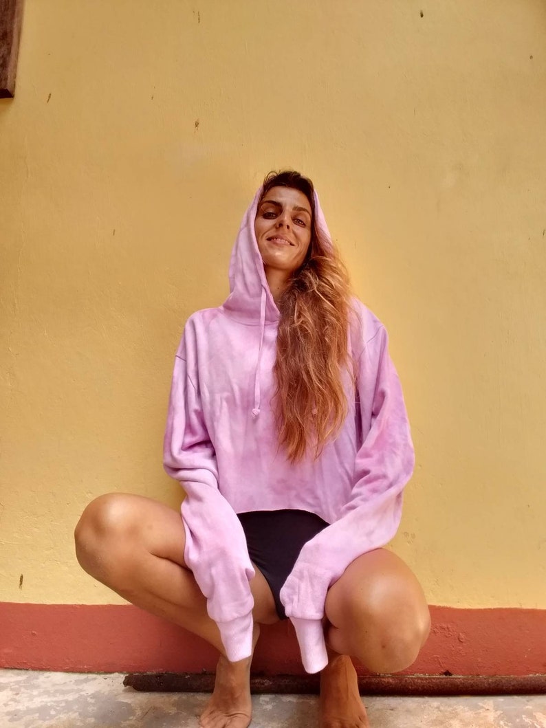 K&ouml;nnte beinhalten: Ein hellrosa Tie-Dye-Hoodie mit Kapuze und Kordelzug sowie langen &Auml;rmeln. Das verk&uuml;rzte Sweatshirt wird mit schwarzen Shorts kombiniert. Das Model hockt vor einer gelben Wand.