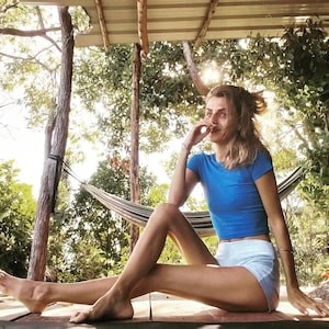 May include: A woman sits on a wooden deck with her legs outstretched. She is wearing a blue crop top and white shorts. She is smiling and looking to the side. There is a hammock behind her.