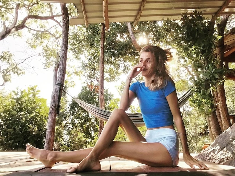 May include: A woman sits on a wooden deck with her legs outstretched. She is wearing a blue crop top and white shorts. She is smiling and looking to the side. There is a hammock behind her.