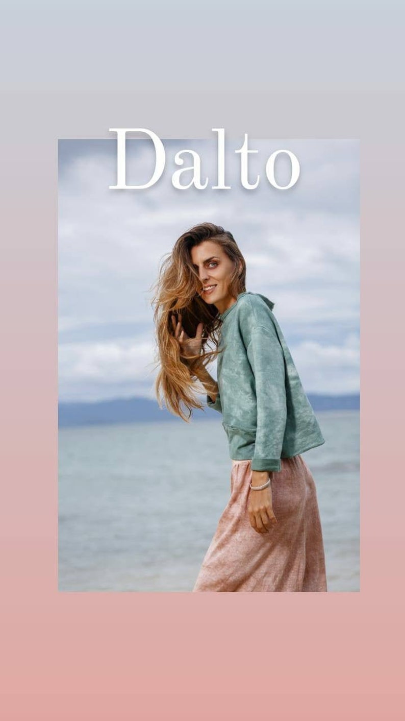 May include: A woman with long brown hair is wearing a light green tie-dye sweatshirt and a light pink skirt. She is standing on a beach with the sea in the background. The text "Dalto" is written in white letters at the top of the image.