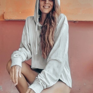 K&ouml;nnte beinhalten: Ein hellgrauer Kapuzenpullover mit langen &Auml;rmeln. Der verk&uuml;rzte Hoodie ist aus einem weichen Material gefertigt. Die Person hockt vor einer roten und orangefarbenen Wand.