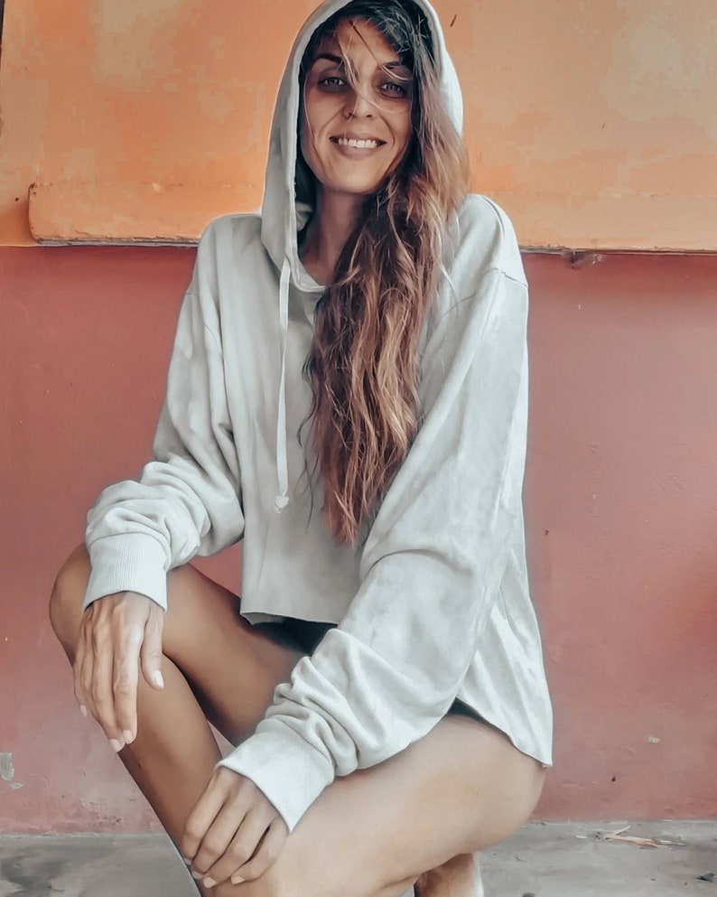 K&ouml;nnte beinhalten: Ein hellgrauer Kapuzenpullover mit langen &Auml;rmeln. Der verk&uuml;rzte Hoodie ist aus einem weichen Material gefertigt. Die Person hockt vor einer roten und orangefarbenen Wand.