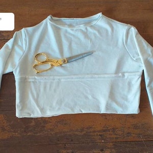 May include: A light blue long-sleeved shirt made of bamboo fleece, laid flat on a wooden surface. The shirt has a round neckline and is ready to be dyed. A pair of gold scissors is placed on top of the shirt.