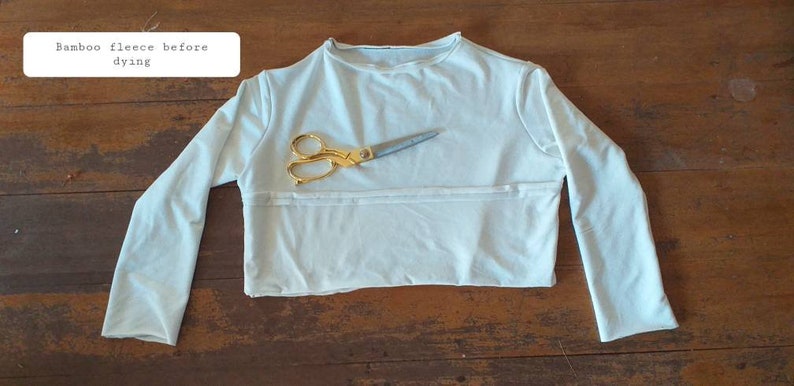 May include: A light blue long-sleeved shirt made of bamboo fleece, laid flat on a wooden surface. The shirt has a round neckline and is ready to be dyed. A pair of gold scissors is placed on top of the shirt.