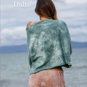 May include: A woman wearing a green tie-dye cropped top and pink tie-dye trousers stands facing away from the camera. The woman is standing on a beach with the sea in the background. The text "Dalto" is in the top left corner of the image.