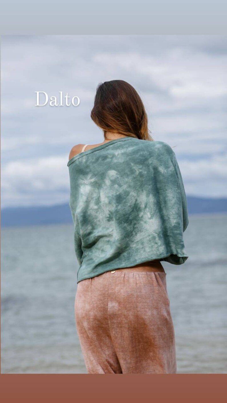 May include: A woman wearing a green tie-dye cropped top and pink tie-dye trousers stands facing away from the camera. The woman is standing on a beach with the sea in the background. The text "Dalto" is in the top left corner of the image.