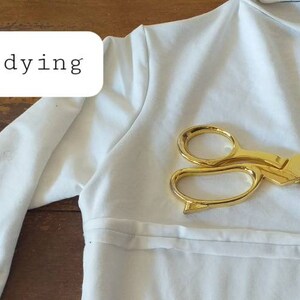 May include: A white long-sleeved shirt with a pair of gold scissors on top of it. The text "Before dying" is written on a white rectangle in the top left corner of the image.