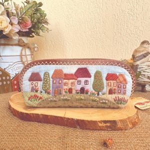 Puede incluir: Un estuche rectangular bordado con una pintoresca escena de pueblo. El diseño incluye casas coloridas con tejados rojos, morados y naranjas, árboles verdes y un campo de flores. El estuche está bordeado con un ribete cosido marrón.