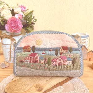 May include: A semi-circular fabric pouch featuring a quilted landscape scene with embroidered houses, trees, and a sun over a body of water. The design uses soft pink, green, and blue tones with detailed stitching throughout the textile surface.