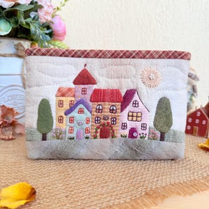 Puede incluir: Una bolsa acolchada con una escena de pueblo bordada. El diseño incluye casas coloridas, árboles y un sol. Las casas tienen tejados rojos, naranjas, morados y rosas. La bolsa tiene un fondo beige con un ribete a cuadros rojos.