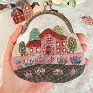 May include: A small, semi-circular coin purse featuring an intricate fabric embroidery of a house with a red roof, surrounded by green trees and colorful flower beds. The purse has a bronze-toned metal kiss-lock clasp closure at the top.