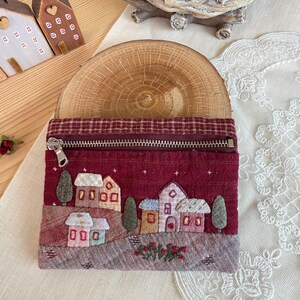 Könnte beinhalten: Eine rechteckige Stofftasche in tiefem Burgunderrot mit gesteppter Textur und einem Metallreißverschluss. Die Vorderseite zeigt eine gestickte Dorfszene mit kleinen Häusern, Bäumen und winzigen Sternen auf dunkelrotem Grund.