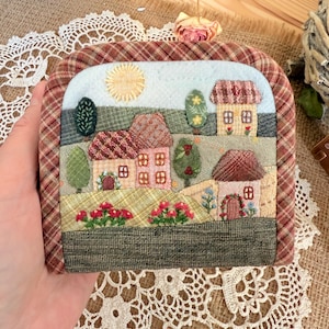 May include: A handmade, quilted landscape scene featuring houses, trees, and a sun. The artwork is framed with a brown and red plaid fabric. The scene is a charming, miniature depiction of a village.