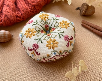 May include: A round, cream-colored pincushion with a floral cross-stitch design in yellow, green, and burgundy. The pincushion has a button in the center and is surrounded by a knitted orange hat, an acorn, and cinnamon sticks.