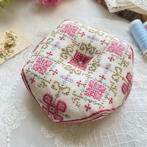 Peut inclure: Un coussin à épingles en tissu blanc avec un motif floral dans les tons de rose, bleu et vert. Le coussin est en forme d'hexagone et a un bouton au centre.