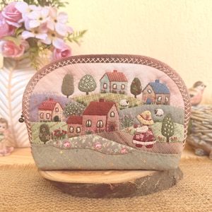 Puede incluir: Un estuche de tela semicircular con bordados detallados de un paisaje rural con varias casas pequeñas, colinas, árboles, ovejas y una figura con sombrero. El diseño utiliza tonos apagados de rosa, verde, marrón y azul.