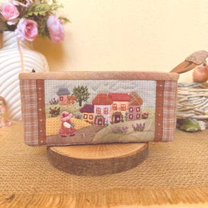 May include: A rectangular fabric pencil case featuring an embroidered scene of a small village with colourful houses, a tree, and a figure in a pink hat walking on a path. The case has a tan plaid border and a zip closure with a fabric pull tab.