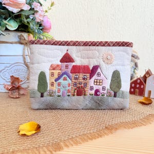 Op de afbeelding: Een gewatteerde stoffen etui met een geborduurde dorpsscène. Het ontwerp omvat kleurrijke huizen, bomen en een zon. De etui heeft een bruin en crèmekleurig kleurenschema met een patroon aan de bovenrand. Het ontwerp is gedetailleerd en grillig.