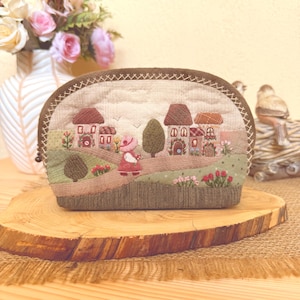 Puede incluir: Un estuche de tela semicircular con una escena de paisaje bordada que incluye casas pequeñas, una figura con vestido rojo, árboles y flores. El estuche tiene un borde marrón oscuro con costuras decorativas y un fondo de tela beige.