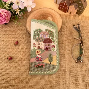 Puede incluir: Un estuche para gafas de tela color verde salvia con una escena bordada de un pequeño pueblo con casas, árboles y una figura con sombrero. El estuche tiene un panel frontal texturizado de color claro con detalles en punto de cruz.