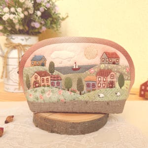 Puede incluir: Un estuche de tela con un paisaje bordado que muestra casas, árboles, un velero en el agua y un sol en el cielo. El diseño incluye motivos florales, colinas verdes y dos ovejas pequeñas. La base del estuche es de tela marrón texturizada.