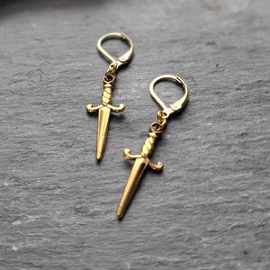 May include: A pair of gold-toned earrings featuring a dagger design. The earrings are made of metal and have a simple, minimalist style.