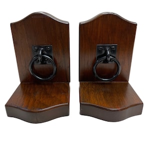 May include: A pair of L-shaped bookends crafted from dark brown stained wood with a smooth finish. Each bookend features a black metal square plate mounted on the vertical panel, holding a circular black metal ring pull centered on the front.