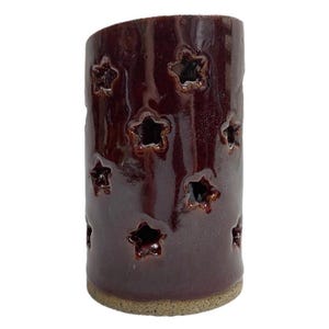 May include: A cylindrical ceramic candle holder with a glossy dark maroon glaze. The exterior surface features multiple star-shaped cutouts arranged in a scattered pattern, allowing light to pass through. The base is unglazed, showing a light tan texture.