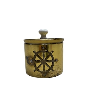 May include: A gold metal jar with a white knob on the lid. The jar has a nautical wheel design on the front.