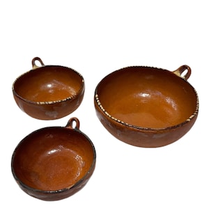 May include: Three brown ceramic bowls with a glossy finish. Each bowl has a small handle and a decorative rim with white accents. The bowls vary in size, suggesting a nesting set.