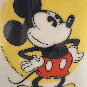 May include: A ceramic mug featuring a graphic of Mickey Mouse standing with hands on hips against a bright yellow circular background. Mickey wears red shorts, white gloves, and yellow shoes. The text WALT DISNEY PRODUCTIONS is printed in black.
