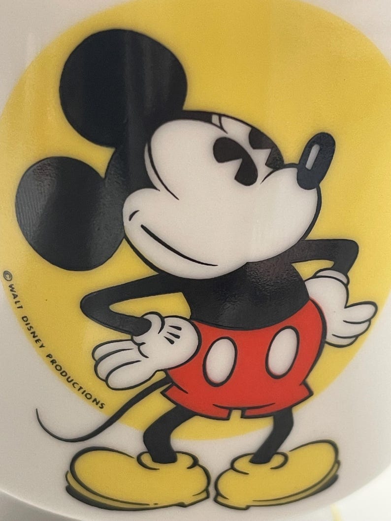 May include: A ceramic mug featuring a graphic of Mickey Mouse standing with hands on hips against a bright yellow circular background. Mickey wears red shorts, white gloves, and yellow shoes. The text WALT DISNEY PRODUCTIONS is printed in black.