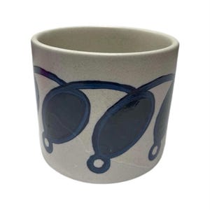 May include: A cylindrical ceramic mug with a speckled off-white base color. The exterior features a repeating hand-painted pattern of dark blue oval shapes connected by thin blue lines, creating a continuous garland-like design around the vessel.