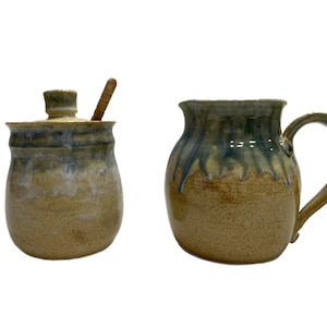 May include: A set of two handmade ceramic honey pots with a rustic glaze. The smaller pot has a lid and a wooden honey dipper. The larger pot has a handle.
