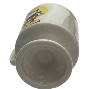 May include: A white ceramic mug with a pedestal base, featuring a graphic of a cartoon mouse with black ears, red shorts, and yellow shoes against a yellow circular background. The mug has a curved handle on the side and a smooth, glossy finish.