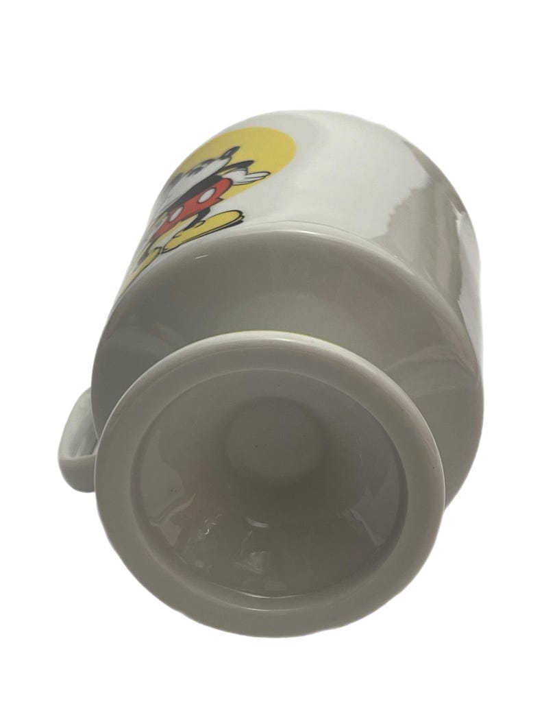 May include: A white ceramic mug with a pedestal base, featuring a graphic of a cartoon mouse with black ears, red shorts, and yellow shoes against a yellow circular background. The mug has a curved handle on the side and a smooth, glossy finish.